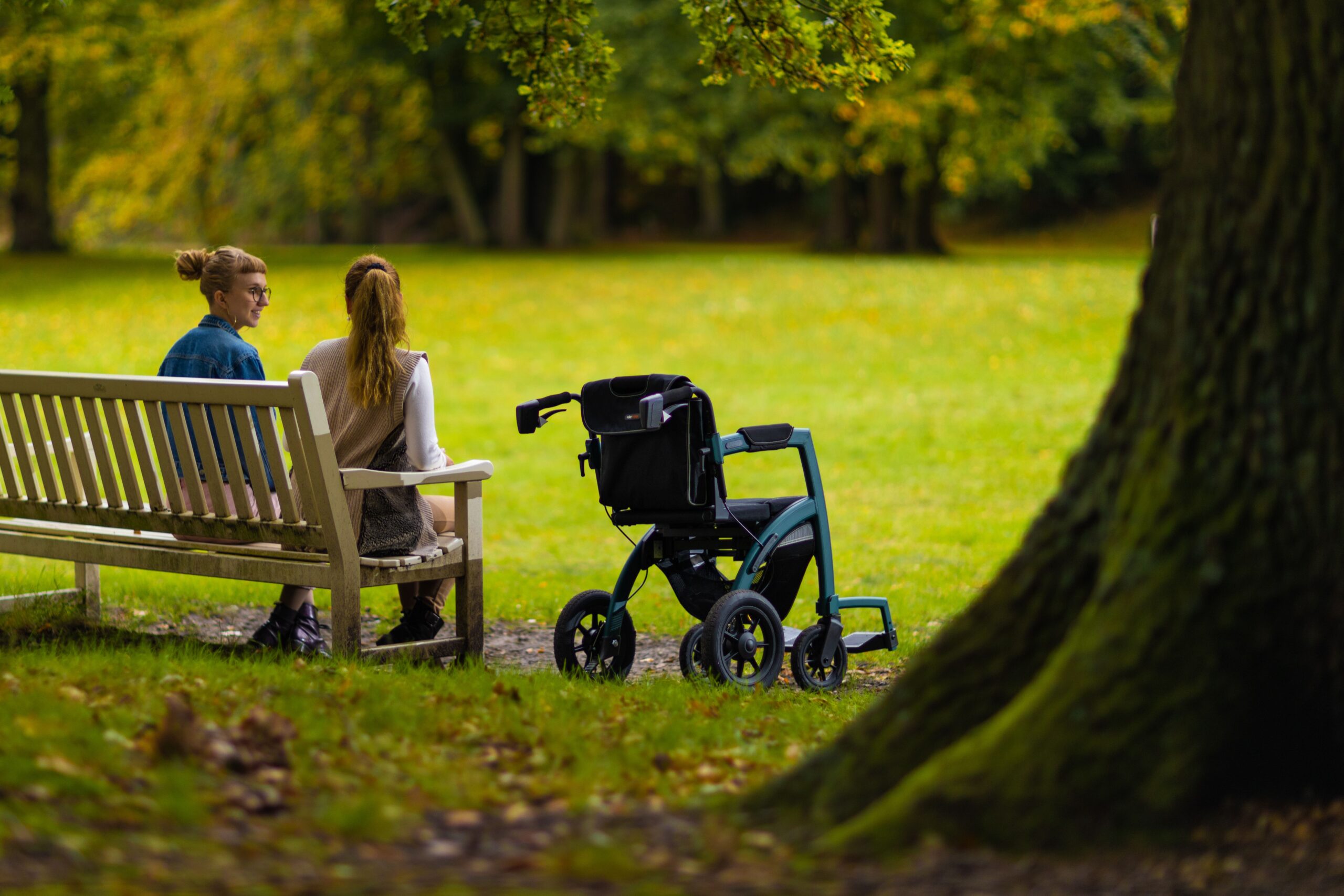 Two women sitting on a bench in a park beside a wheelchair, enjoying a peaceful day outdoors.